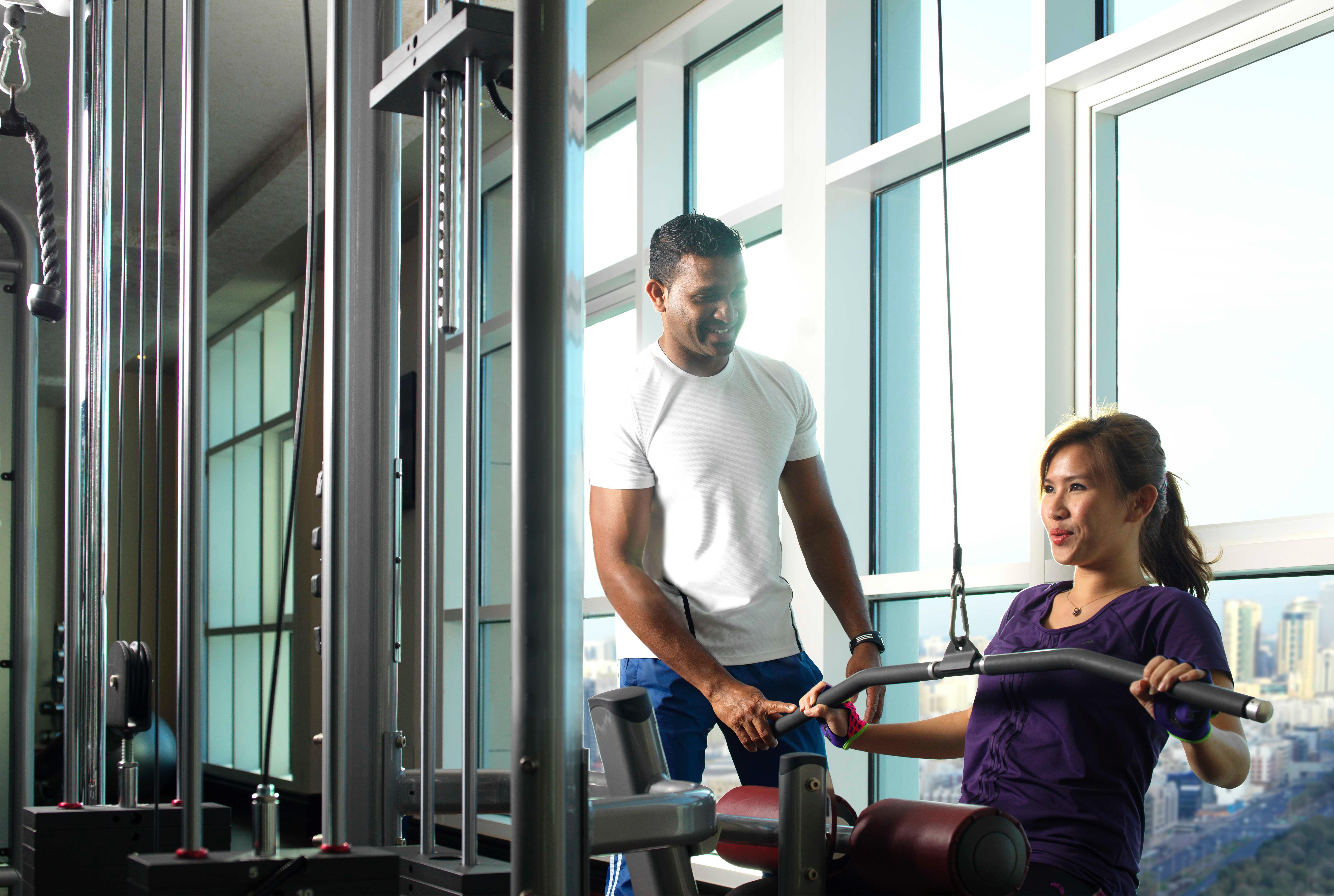 Dusit Thani Abu Dhabi Hotel Exterior photo The photo shows two individuals in a gym setting. One person, likely a trainer, is assisting a woman who appears to be exercising on a machine. The gym has large windows that let in natural light, creating a bright atmosphere. The trainer is focused 